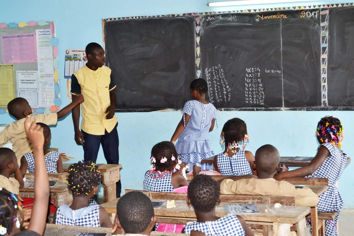 Une classe primaire à Abidjan : les élèves en uniforme kaki et robe vichy bleue, des mains levées, une élève au tableau noir devant l'instituteur